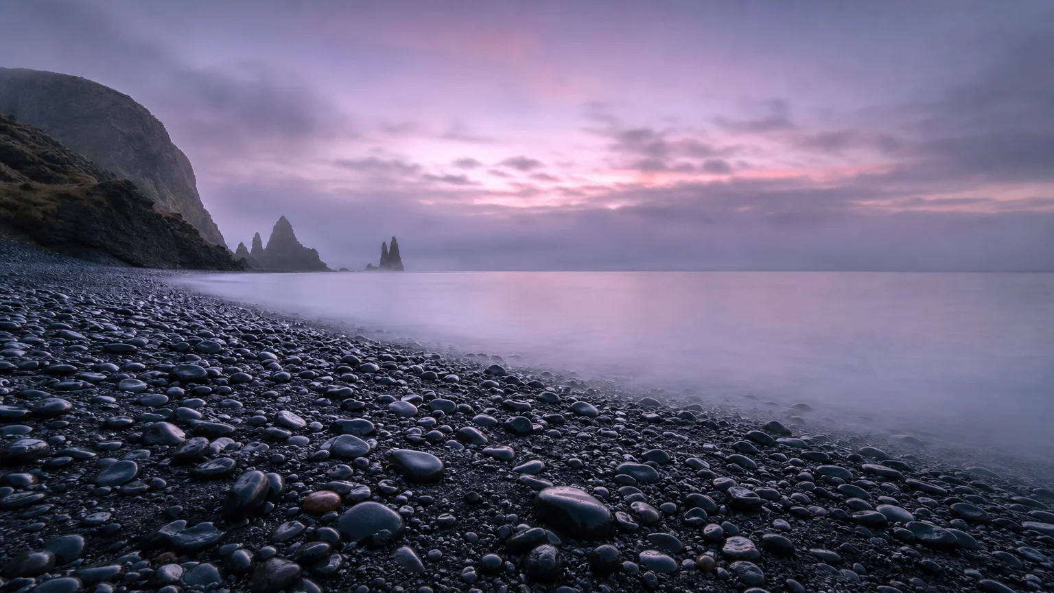 A wide, misty photograph of a calm, dark pebble beach and ocean at dawn, symbolizing environmental preservation.
